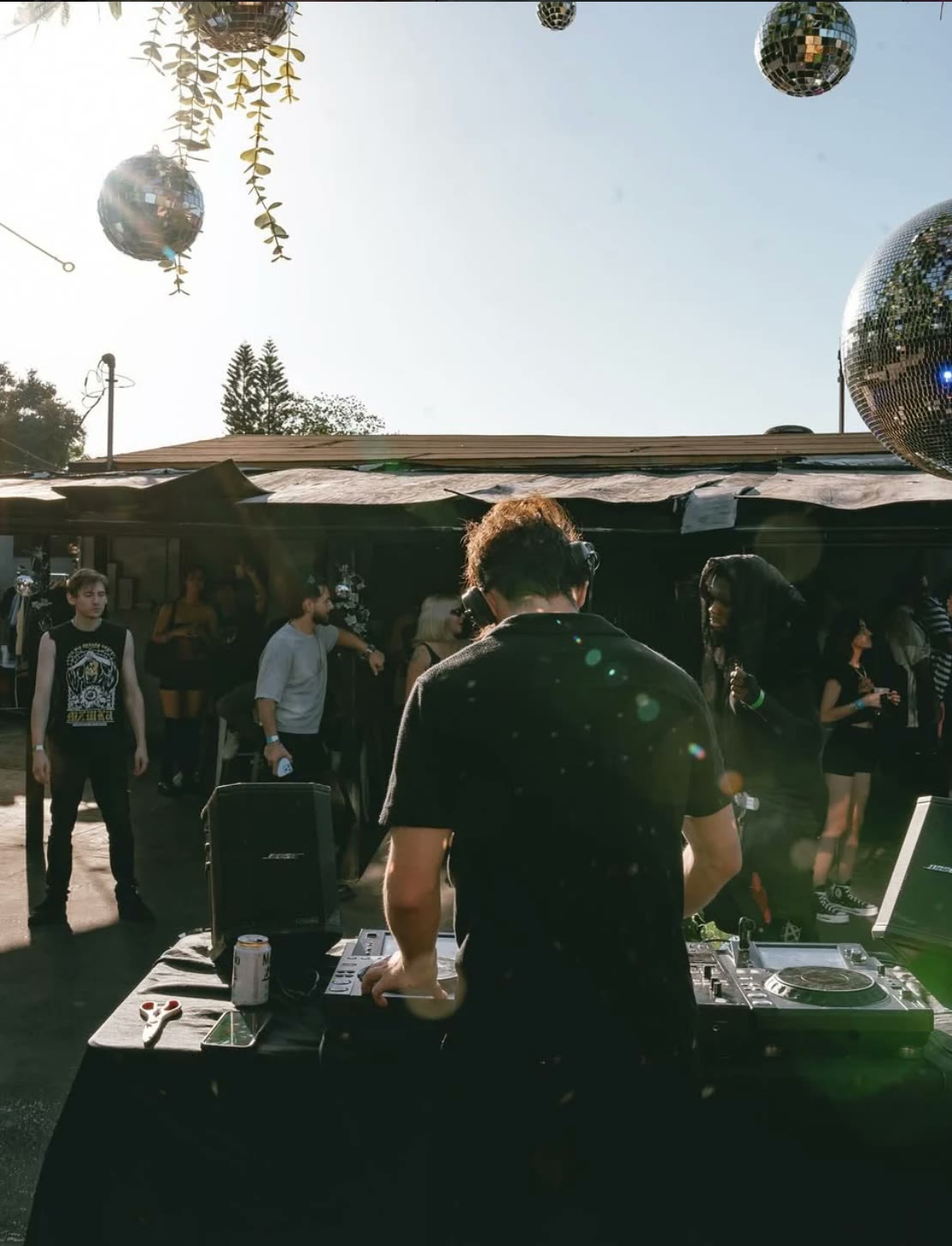 Ryan R playing an outdoor daytime set under hanging disco balls, the crowd behind him
